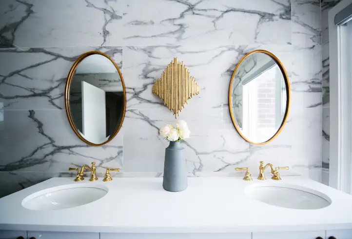 Modern bathroom with a marble wall, gold-framed oval mirrors, gold accents, a vase with white flowers, and a bright doorway reflection
